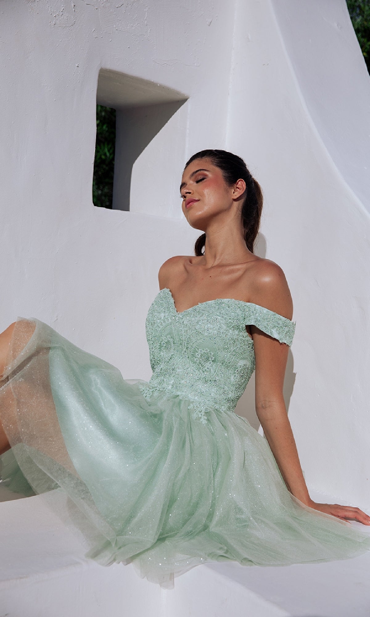 A woman in the light green Off-the-Shoulder Short Hoco Dress: Eureka 9833 sits against a white textured wall with geometric cutouts, looking slightly upward with her eyes closed.